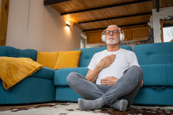 Man with withe beard sitting on the floor against the couch with headset on, eyes closed, hand on heart practicing meditation or mindfulness
